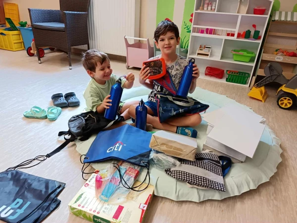 Két mosolygó fiú a szőnyegen, ajándékba kapott kulacsokkal, tornazsákokkal és rajzlapokkal